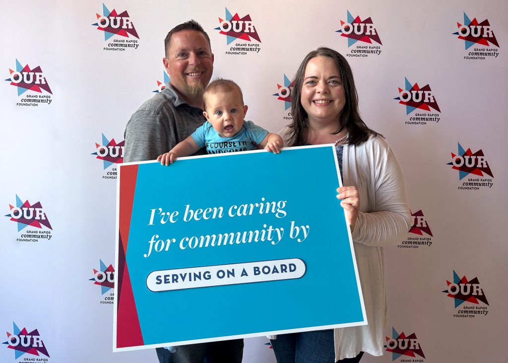 Two adults and a baby hold a large sign that reads “I’ve been caring for community by serving on a board” in front of a Grand Rapids Community Foundation step-and-repeat backdrop.