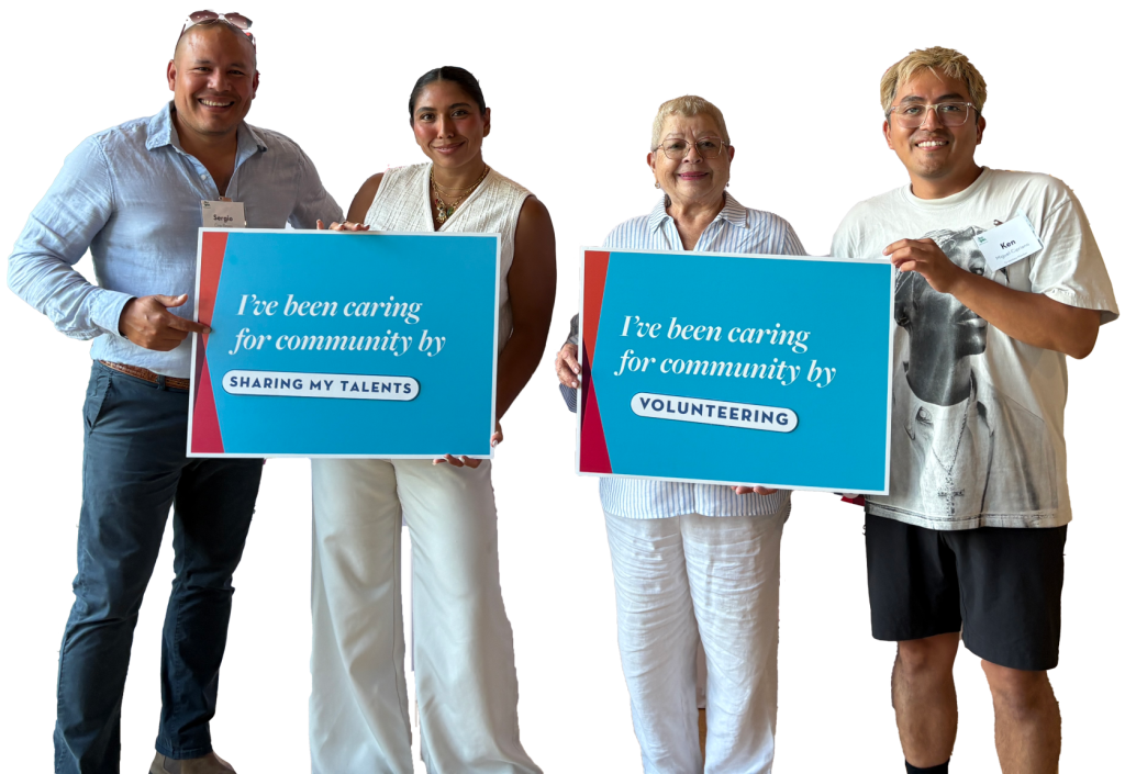 Four people standing together holding blue signs that read ‘I’ve been caring for community by sharing my talents’ and ‘I’ve been caring for community by volunteering’ in front of a colorful geometric background.